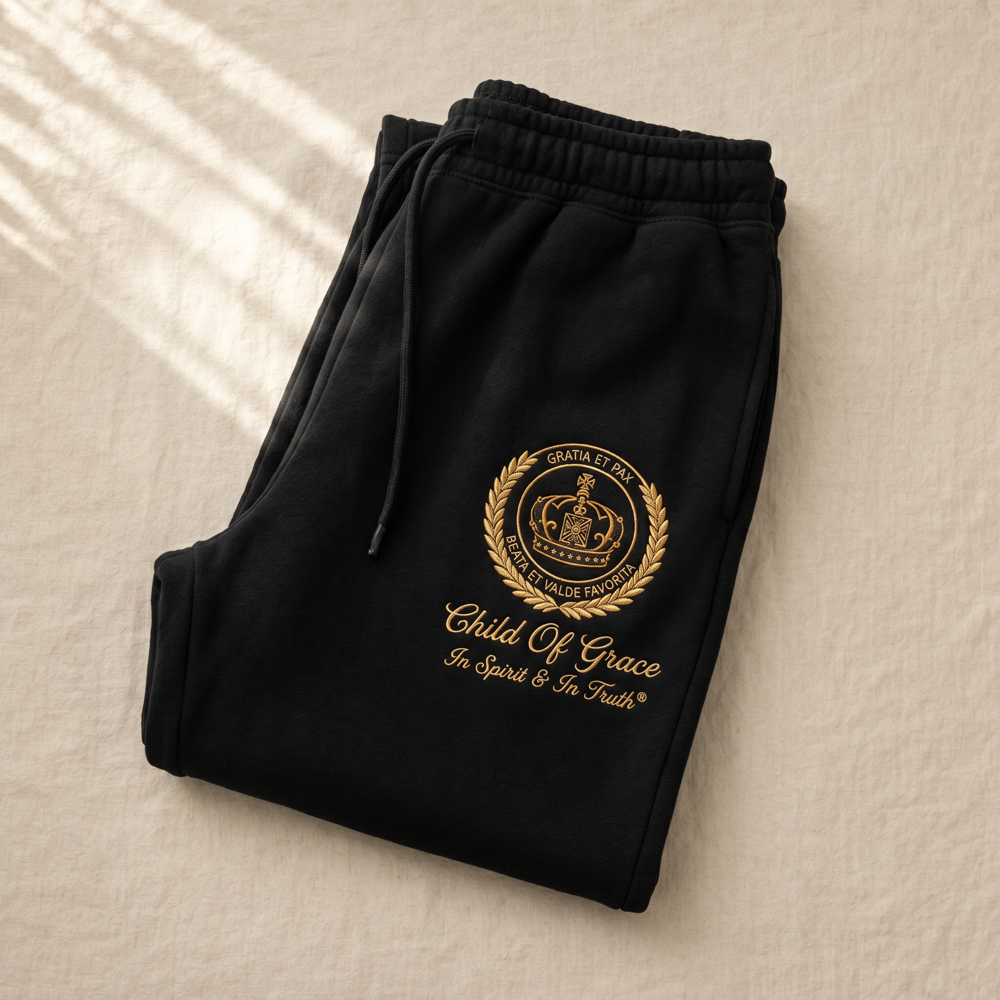 Genesis Crest Sweatpants — Black with antique gold embroidered crest, folded flat-lay on cream linen