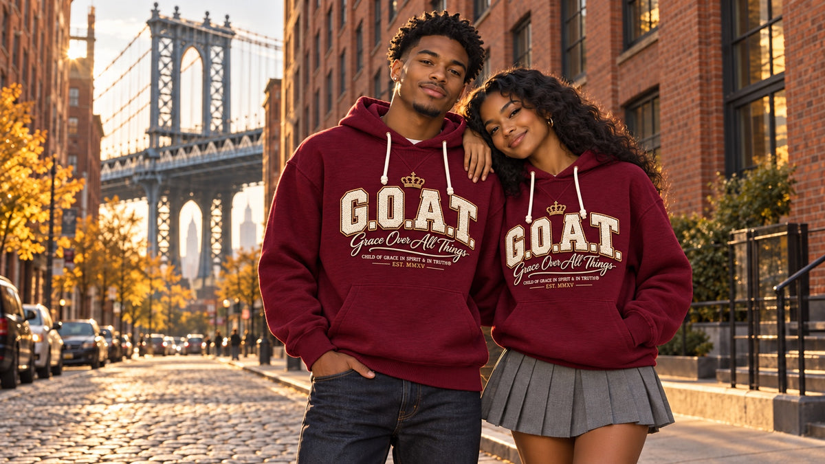 A collegiate-style couple wearing the Child Of Grace burgundy G.O.A.T. Grace Over All Things hoodie in DUMBO, Brooklyn, with the Manhattan Bridge behind them